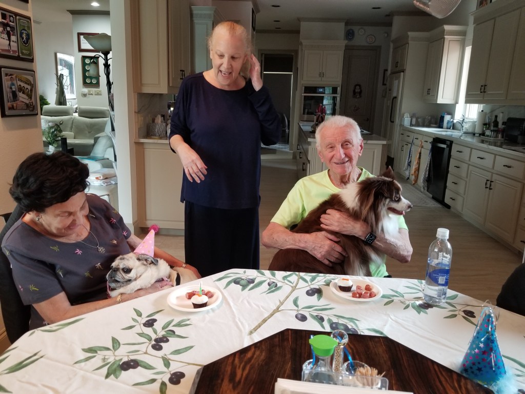 A family photo. We are singing Happy Birthday to Daisy. Claudia’s mom is on the left holding our Pug Daisy. Claudia's dad is on the right holding our mini-Australian Shepherd Rollo. They are both sitting. Claudia is standing up in the middle.