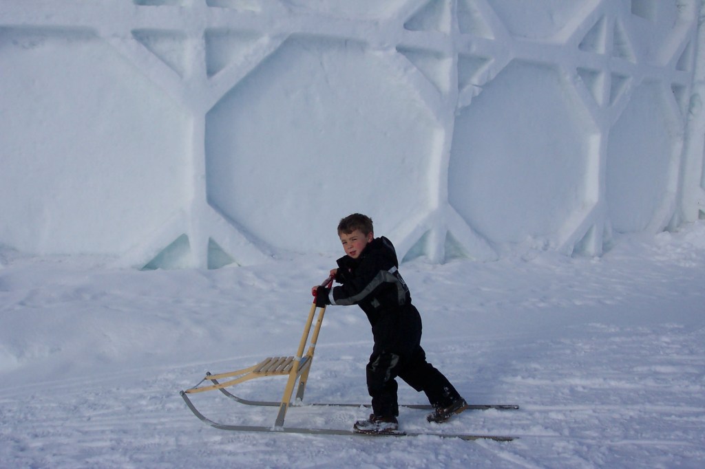 He is standing and kicking with his feet to move forward with the sled. He is passing in front of the ice theater.