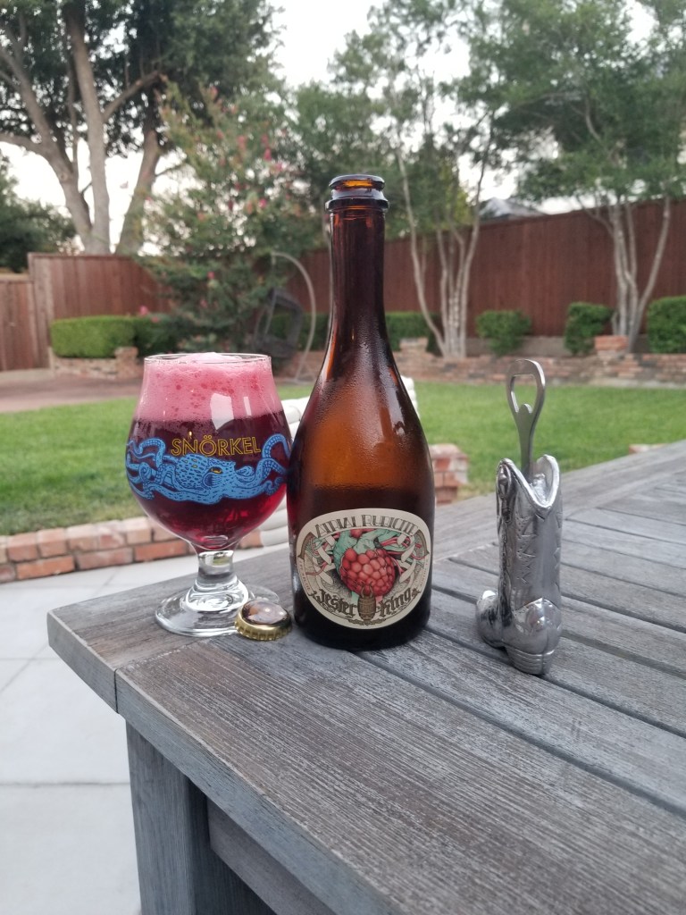 A bottle of Atrial Rubicite a so called Wild Ale from Jester King, Texas, is standing on a table in my backyard. There is a glass filled with Atrial Rubicite and boot shaped opener.