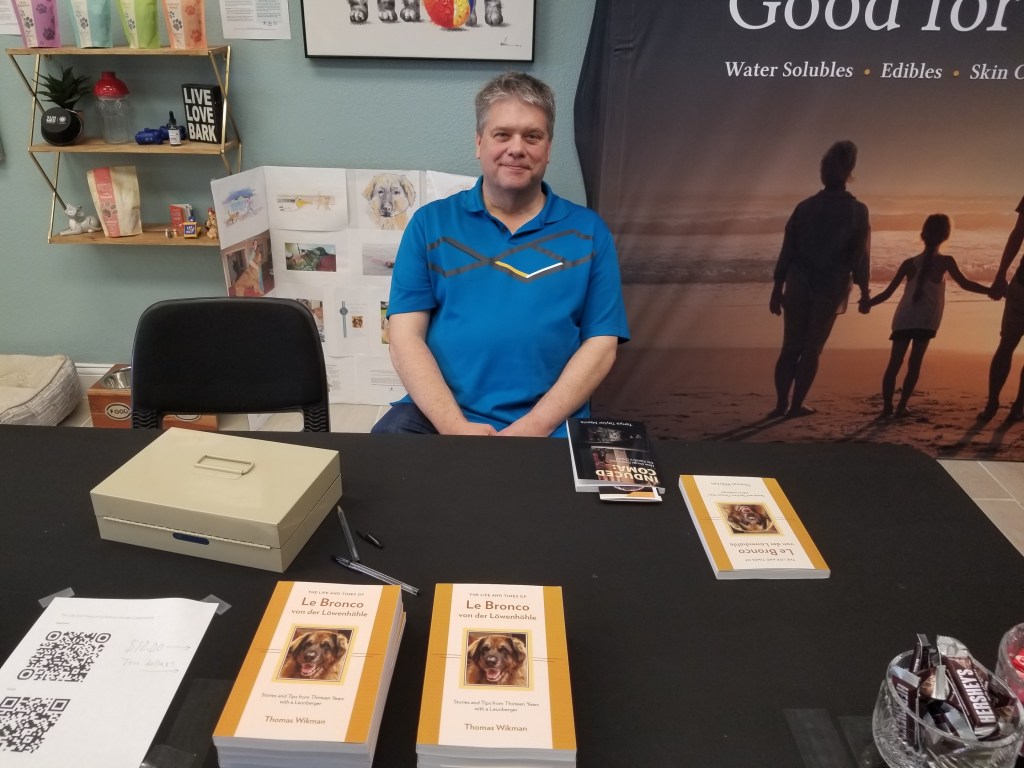 Photo of me sitting at my book signing table at Your CBD Store, Sunmed in Southlake, Texas. I am wearing a blue polo and my table feature several samples of my book, a metal box with money, and a sheet with QR codes.