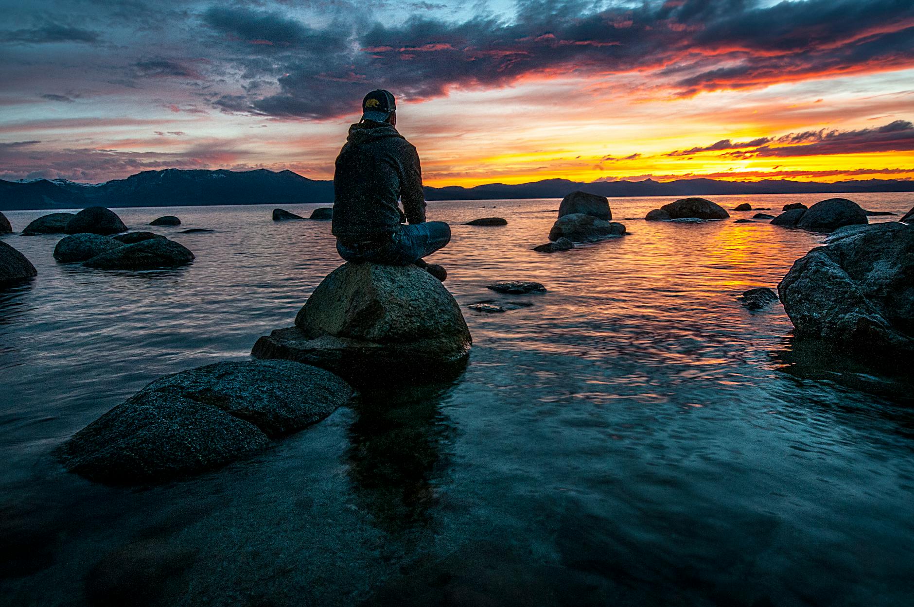 A man sitting on a rock by the ocean look at the senset.