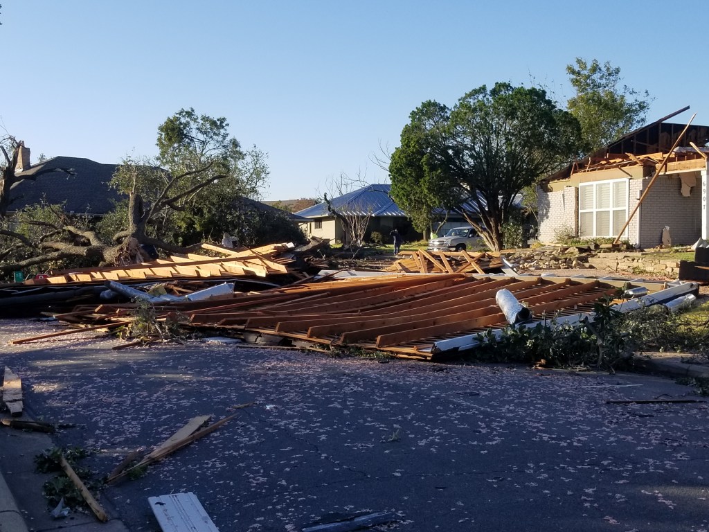 A photo of severely damaged house. The roof is lying in the street.