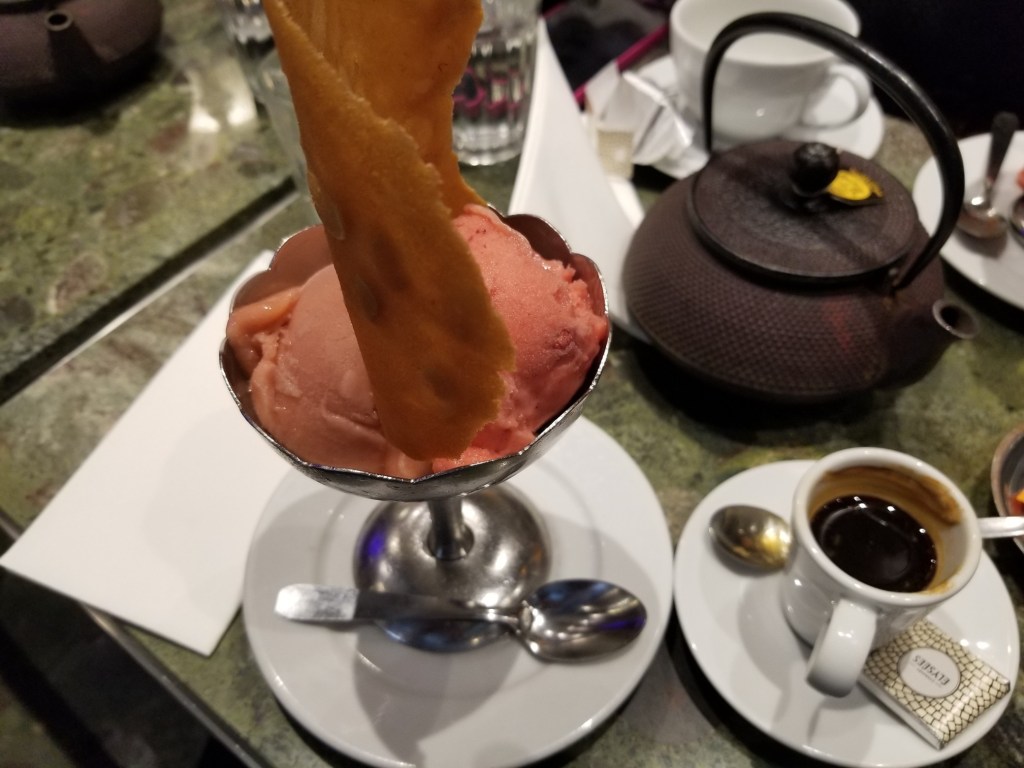 The photo shows rhubarb and wild strawberry ice cream with a big wafer in a silver goblet. There is also a coffee cup and a teapot.