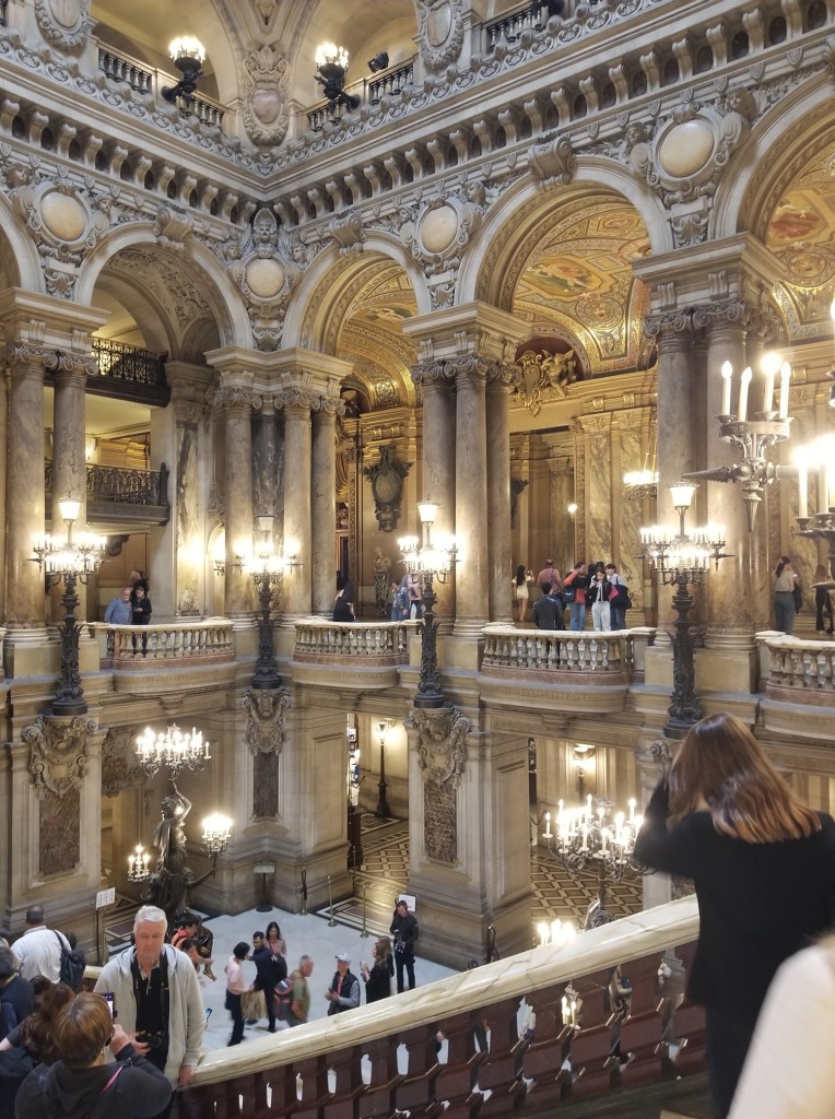 The photo shows a very large room featuring columns, stairs and balconies. It is the second largest room in Palais Opéra Garnier and close to the entrance.