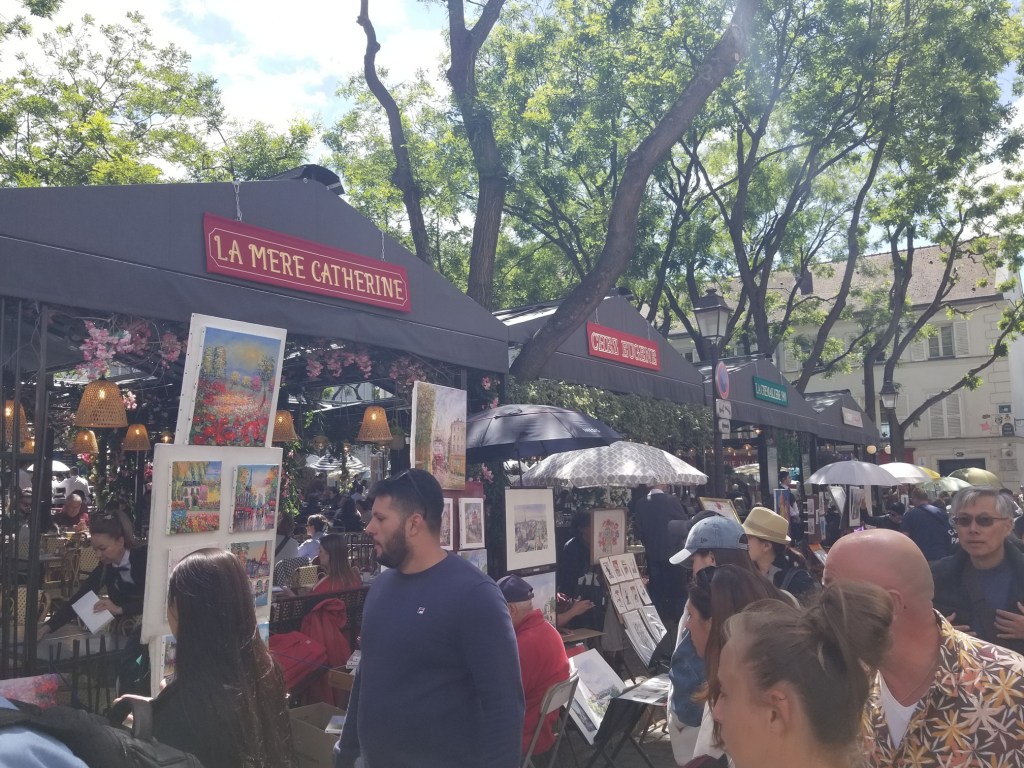 A row of stands where vendors are selling art. There are a lot of people in the photo.