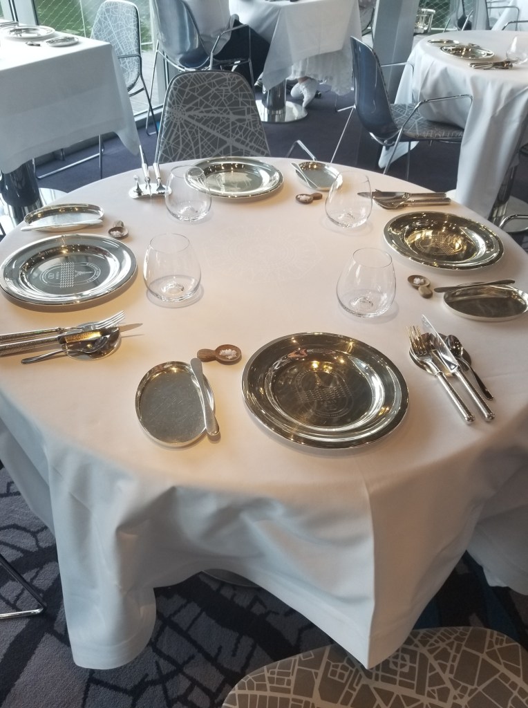 Four silver plates on a table with a white cloth situated in a dining room on a boat.