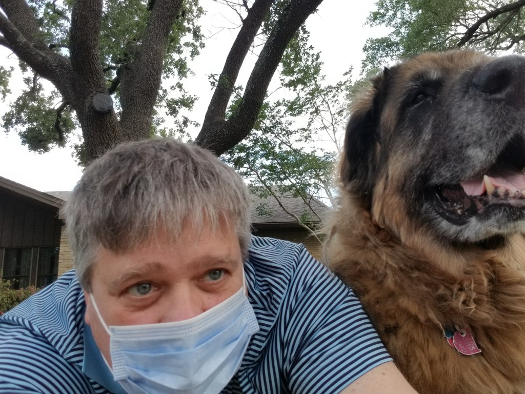 Our Leonberger Bronco and me are lying on the ground looking into the camera. I am wearing a mask. I am wearing a blue polo short.