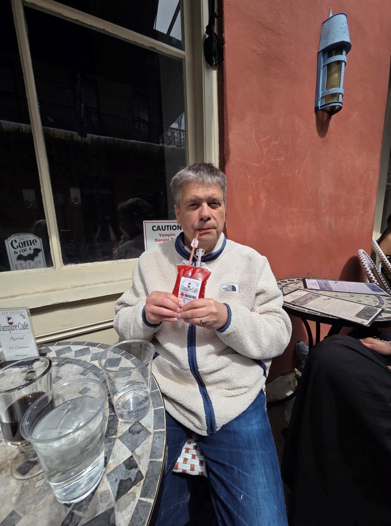 I am sitting on a chair in front of the Vampire Café drinking from a blood bag with a straw.