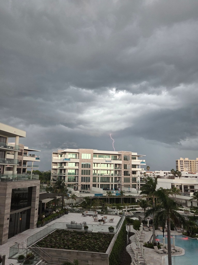 A white and beige six story building with lighting above its roof. It looks like it is striking the building, but the lightning flash is really behind the building.